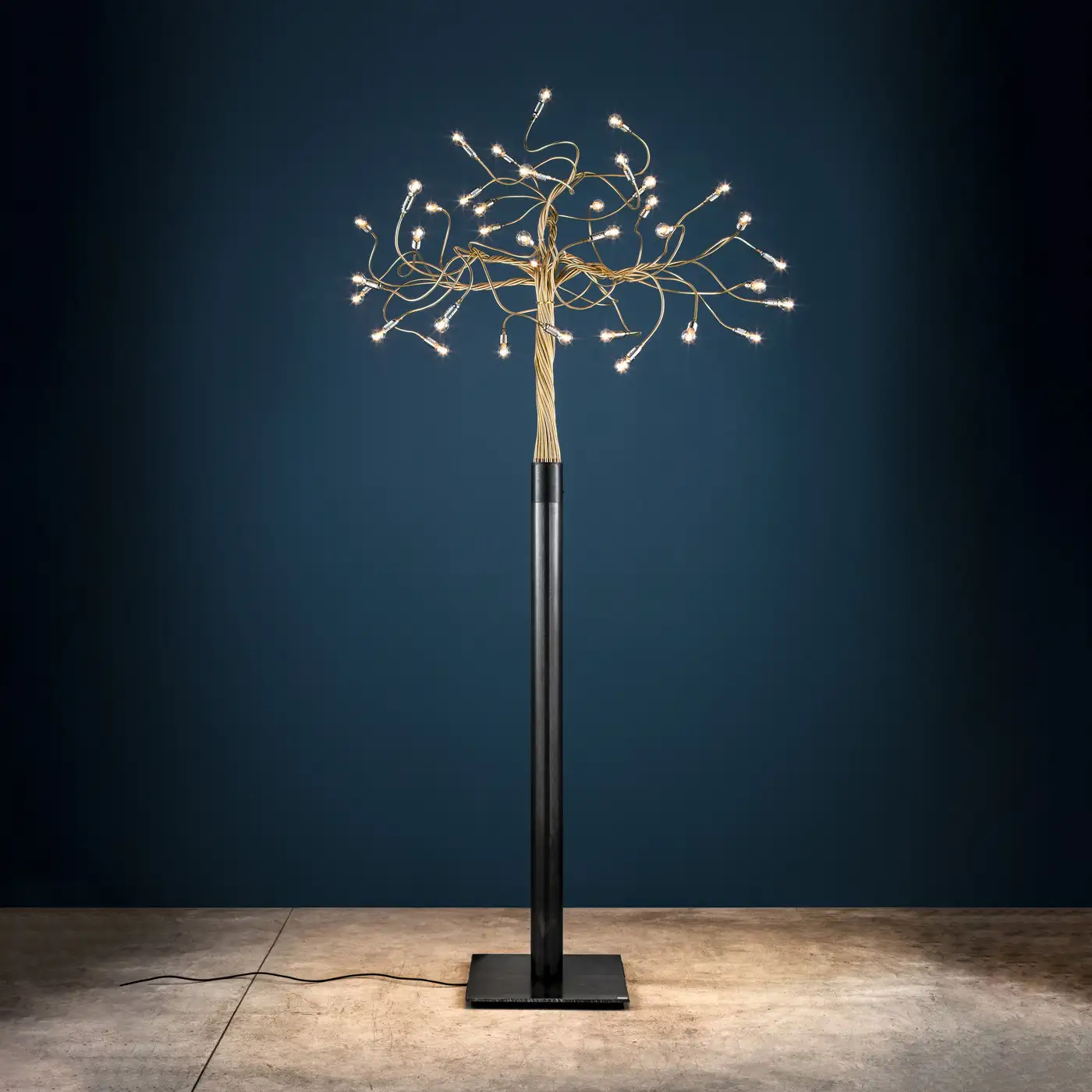 Der Leuchter 'Albero della luce' von Catellani & Smith wirkt wie ein filigraner Baum und steht hier vor einer dunkelblauen Wand.