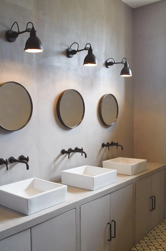 Drei viereckige Waschbecken mit schwarzen Wasserhähnen, über denen jeweils ein runder Spiegel und eine Wandleuchte von DCW hängen