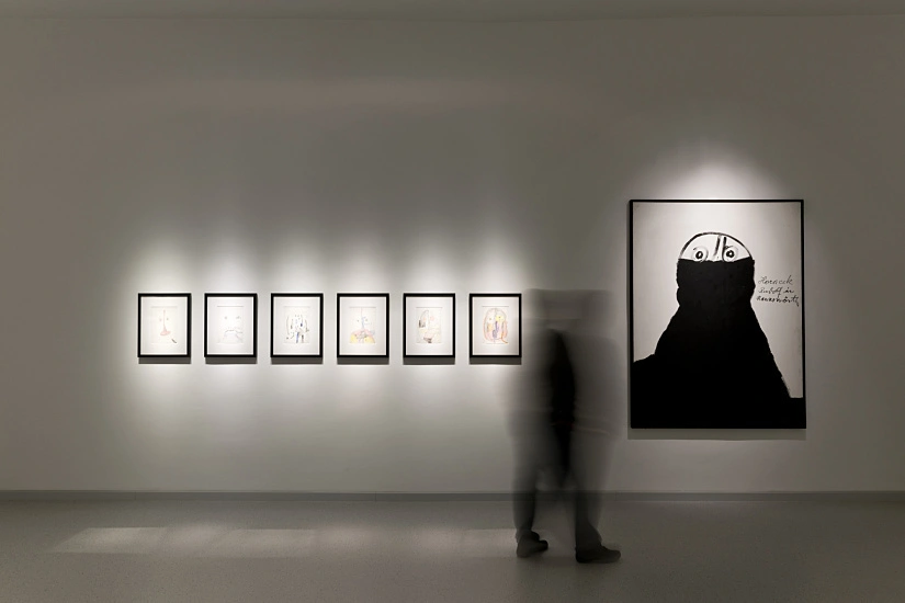 An einer Galeriewand hängen sechs kleine Bilder und eine große Zeichnung, erhellt durch einzeln gesetzte Beleuchtung.