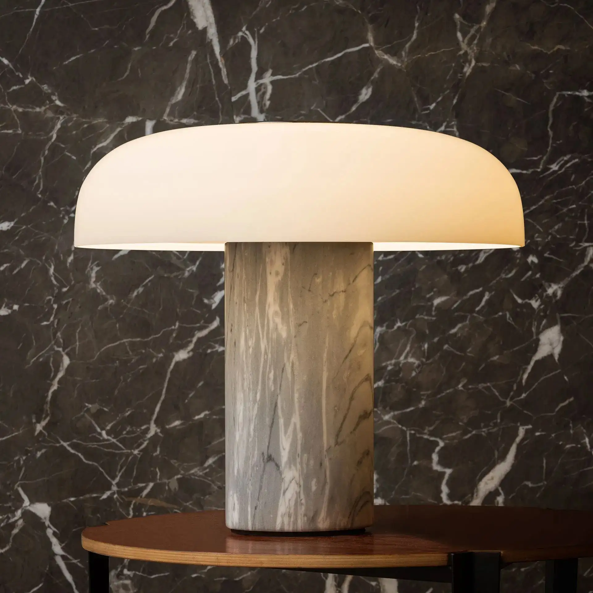 Die Leuchte 'Tropico' von Fontana Arte steht auf einem runden Holztisch vor einer Marmorwand.