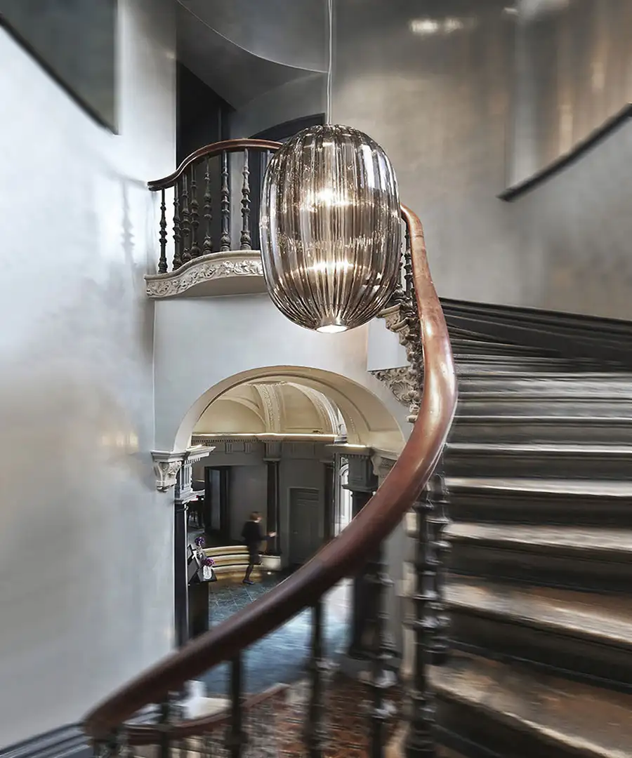 Eine alte Holztreppe in einem klassischen Gebäude; im Treppenauge hängt die Leuchte 'Plass' von Foscarini.