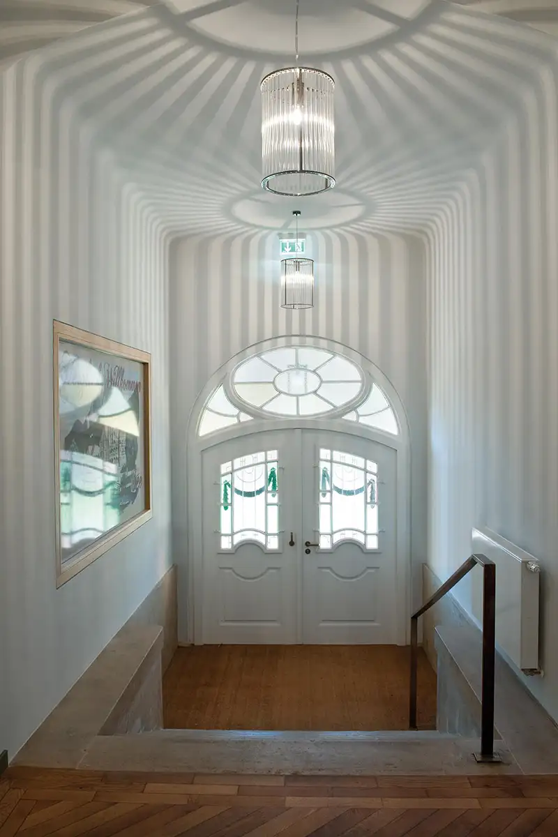 Der Eingangsbereich eines klassizistischen Hauses, erhellt von zwei zylinderförmigen Deckenleuchten, die ein Lichmuster an die Decke und die Wände werfen.