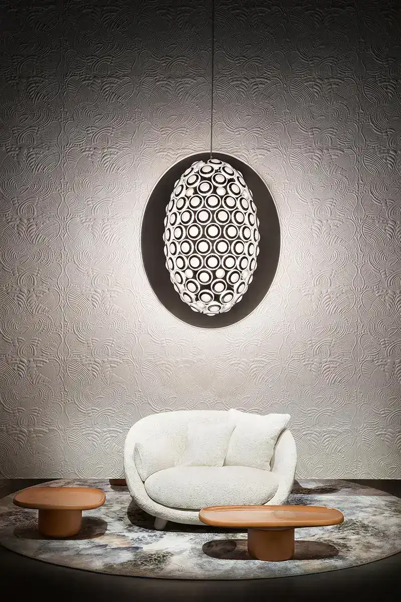Eine große ovale Leuchte hängt über einem kleinen weißen Sofa vor einer strukturierten Wand.