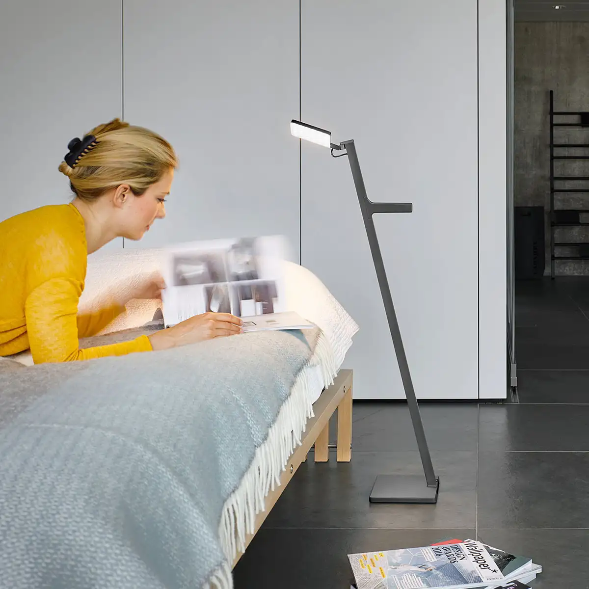 Die Standleuchte 'Roxxane Leggera' von Nimbus steht neben einem Bett und beleuchtet die Zeitschrift, die dort gelesen wird.