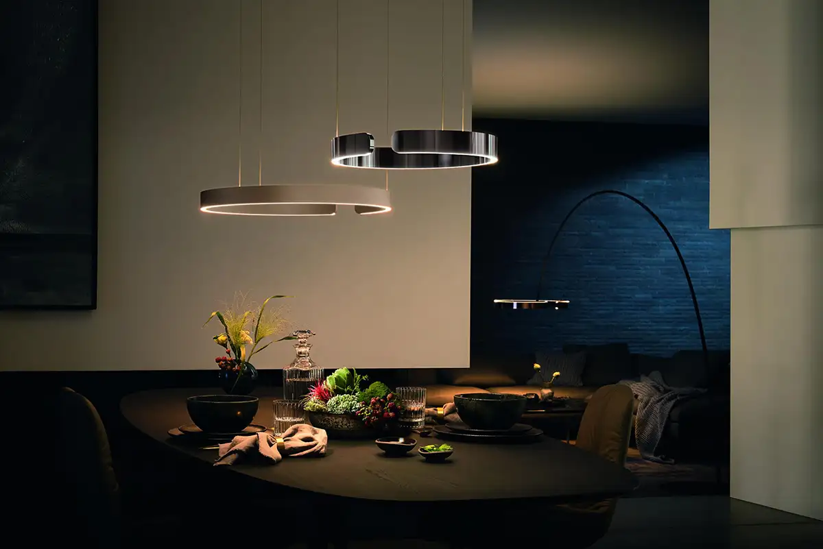 Zwei kreisförmige Leuchten hängen über einem stimmungvollen Stilleben-Arrangement auf einem Esstisch mit dunklem Untergrund.