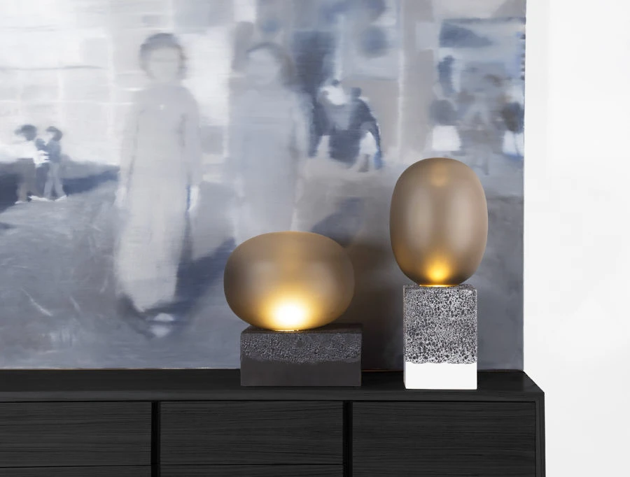Zwei Leuchten 'Magma' von Pulpo aus rauchgrauem Glas und Stein auf einem Sideboard vor einem Gemälde.