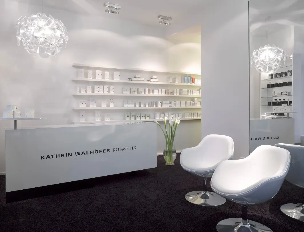 Blick in das Entrée eines Kosmetikinstituts mit weißen Wänden und weißen Möbeln auf dunklem Boden.